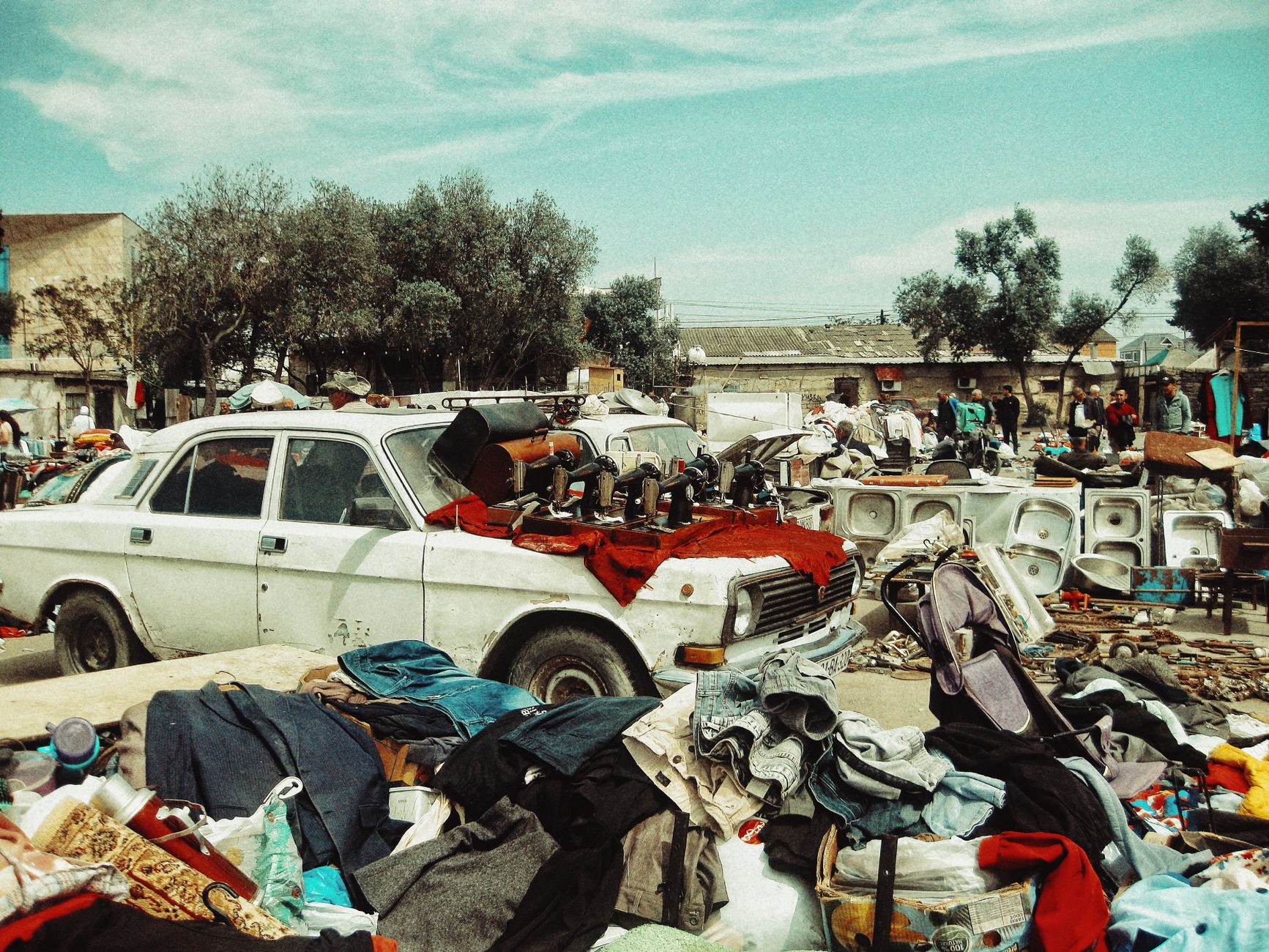 A vintage car overloaded with items at a bustling outdoor flea market under clear skies.