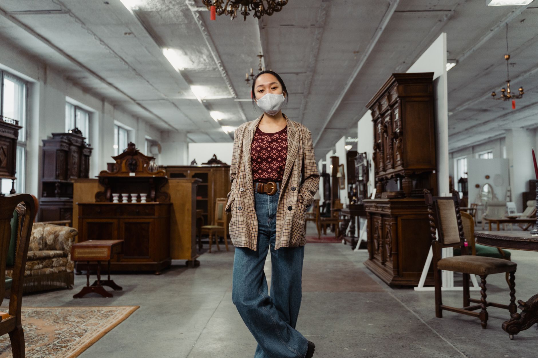 A woman wearing a mask stands inside an antique store surrounded by vintage wooden furniture.