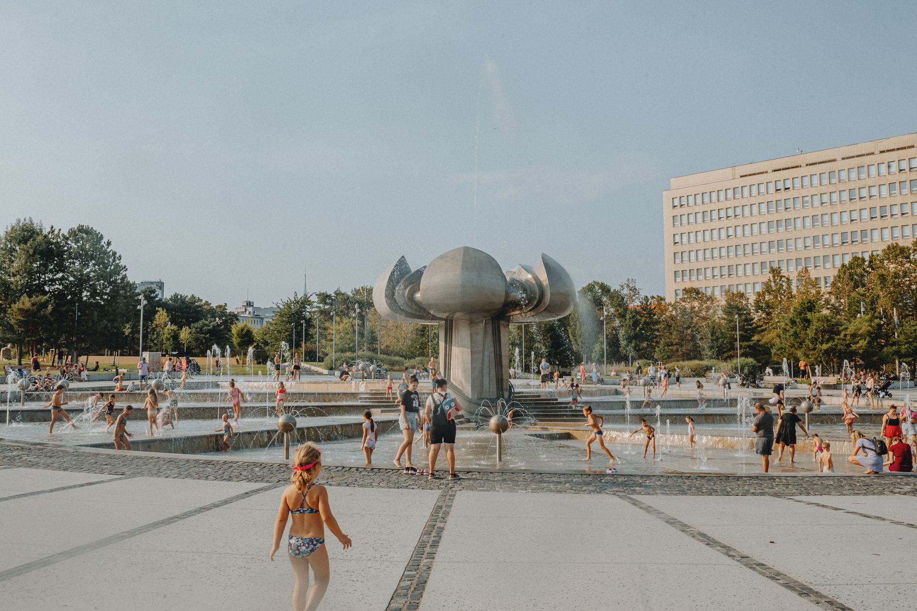 Children enjoying a sunny day at an urban park fountain with modern architecture.