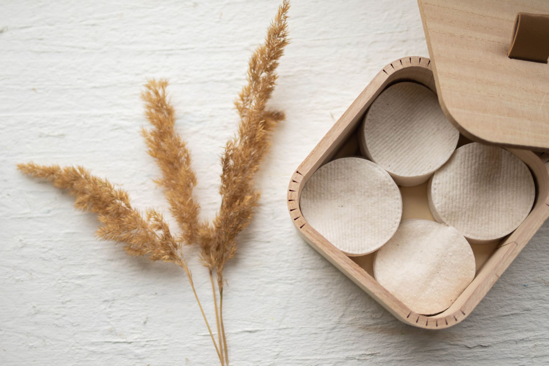 Minimalist still life of a wooden box with eco-friendly cosmetic pads and dried grass on a white background.