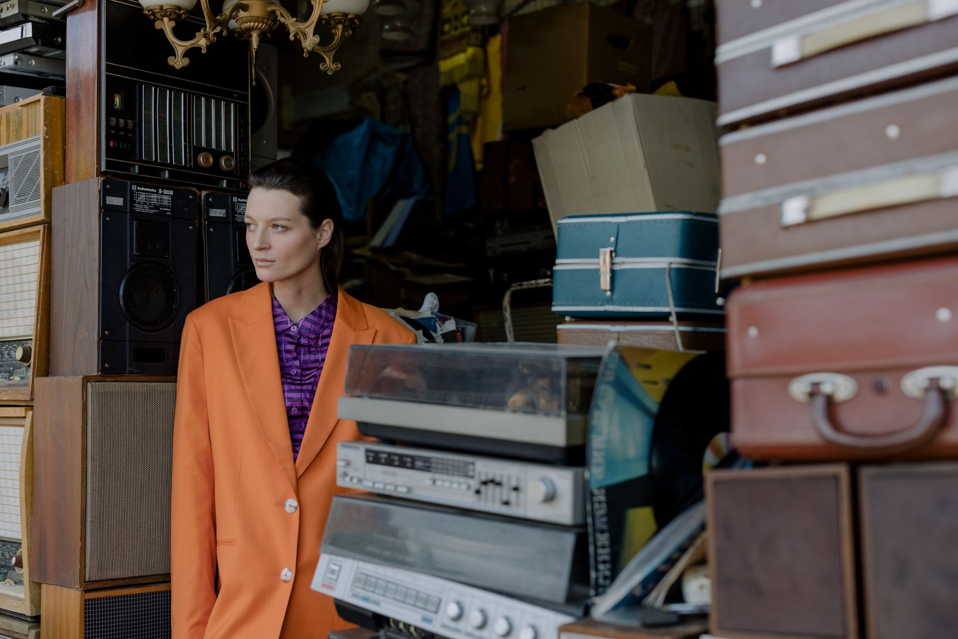 A woman in an orange jacket stands among vintage electronics and suitcases in an eclectic shop.