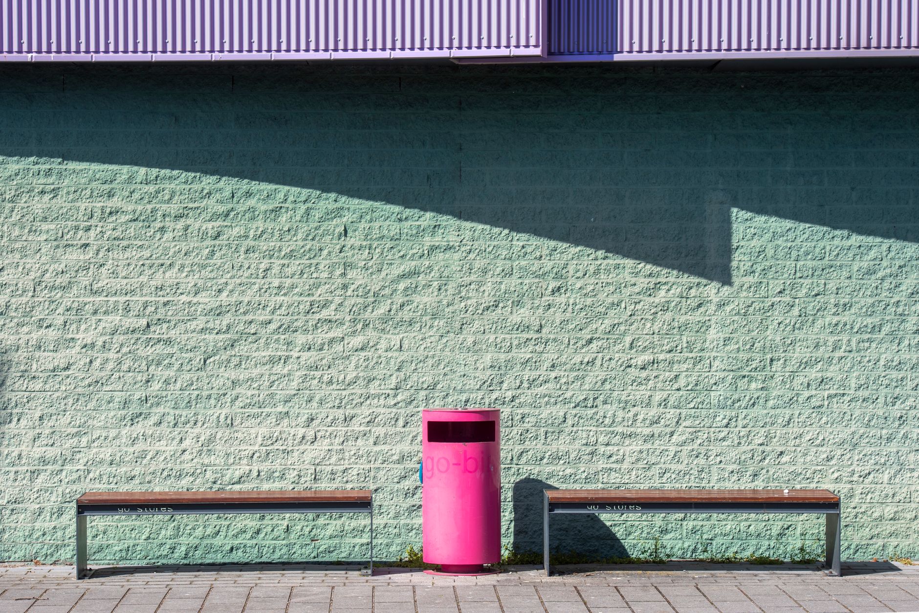 A minimalist urban scene featuring benches and a bright pink trash bin against a colorful wall.