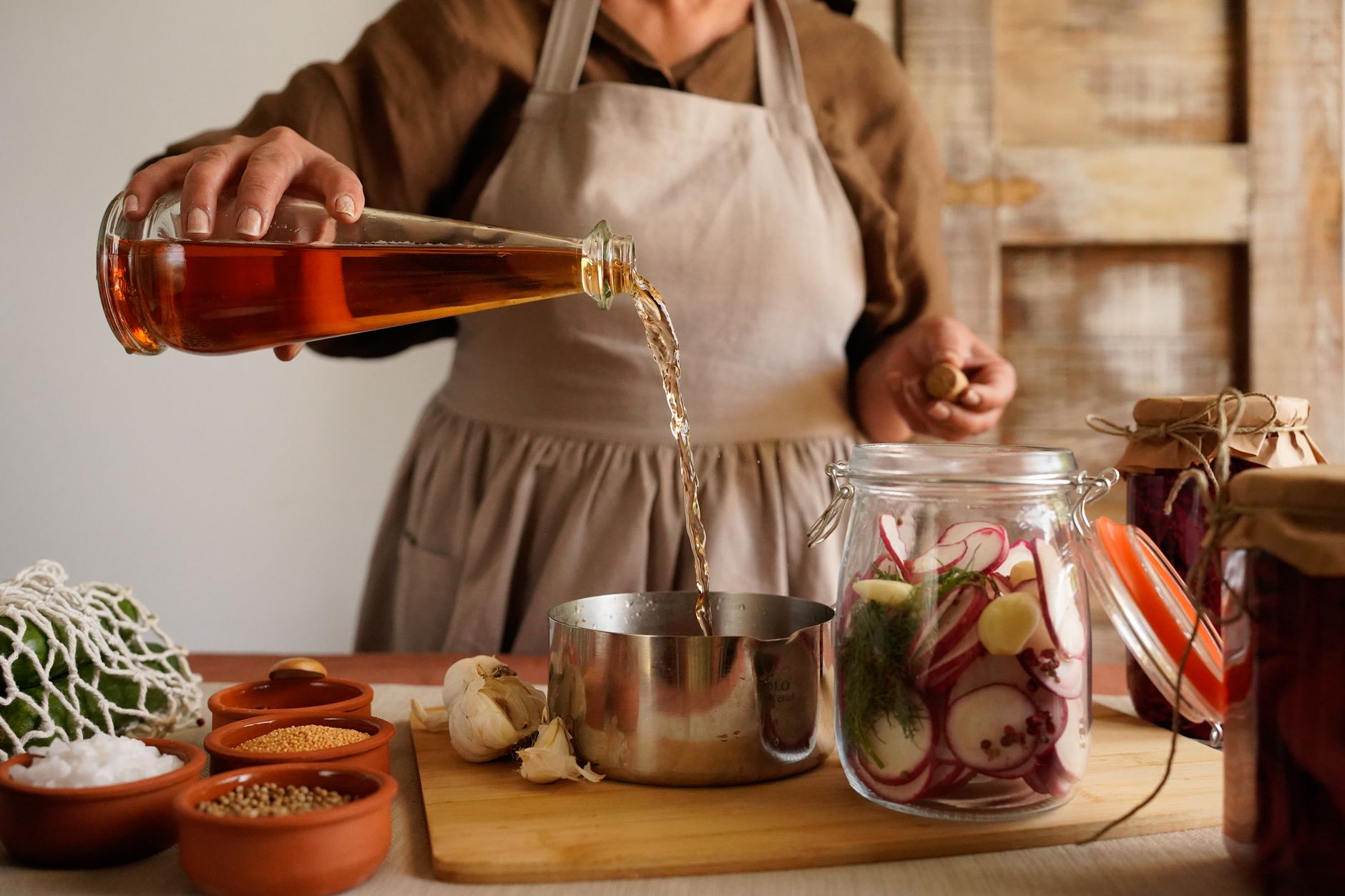 A woman pours vinegar into a pot while making homemade pickles in a rustic kitchen setting.
