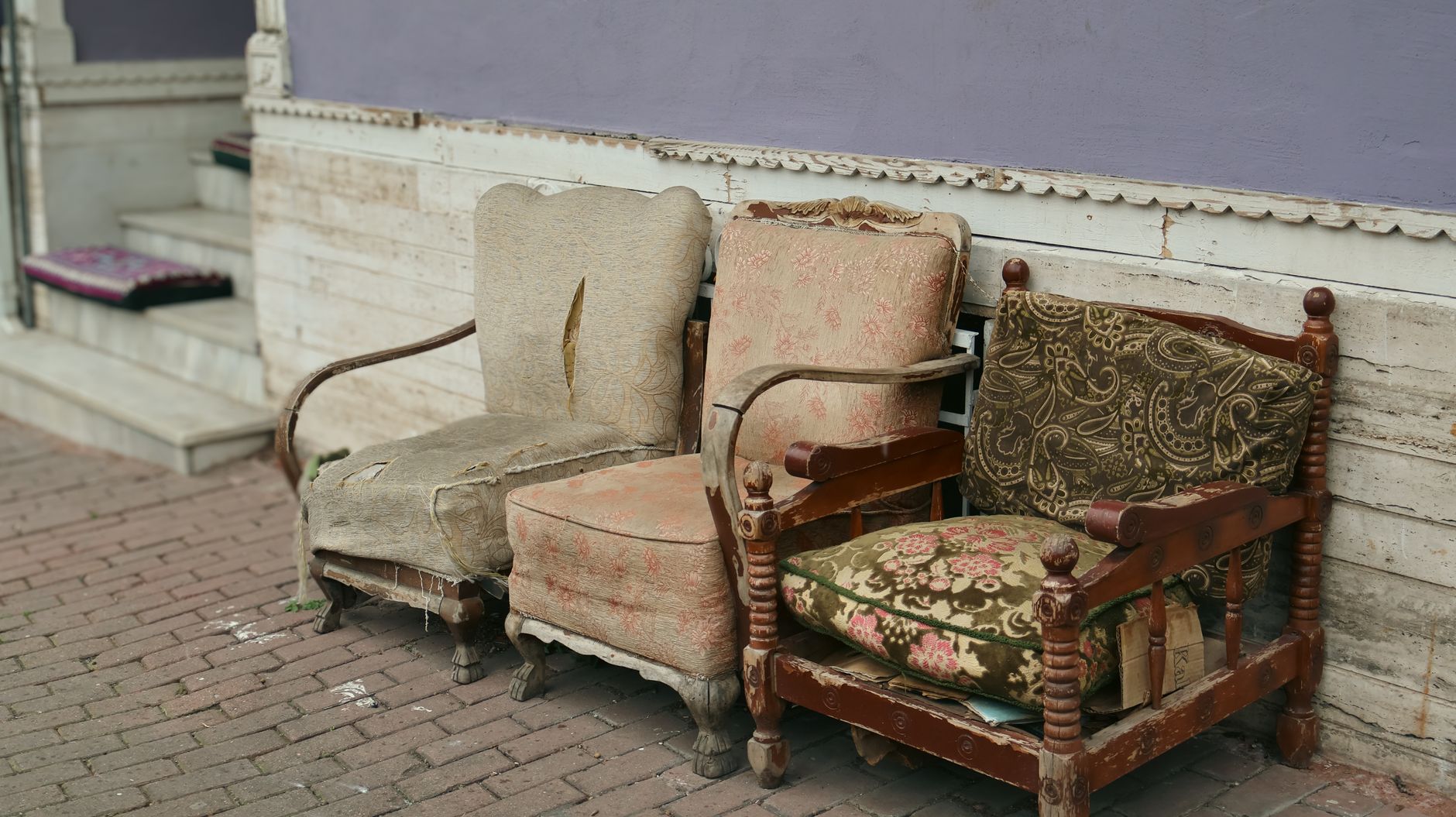 Three vintage chairs with worn upholstery sit outside on a rustic brick sidewalk.