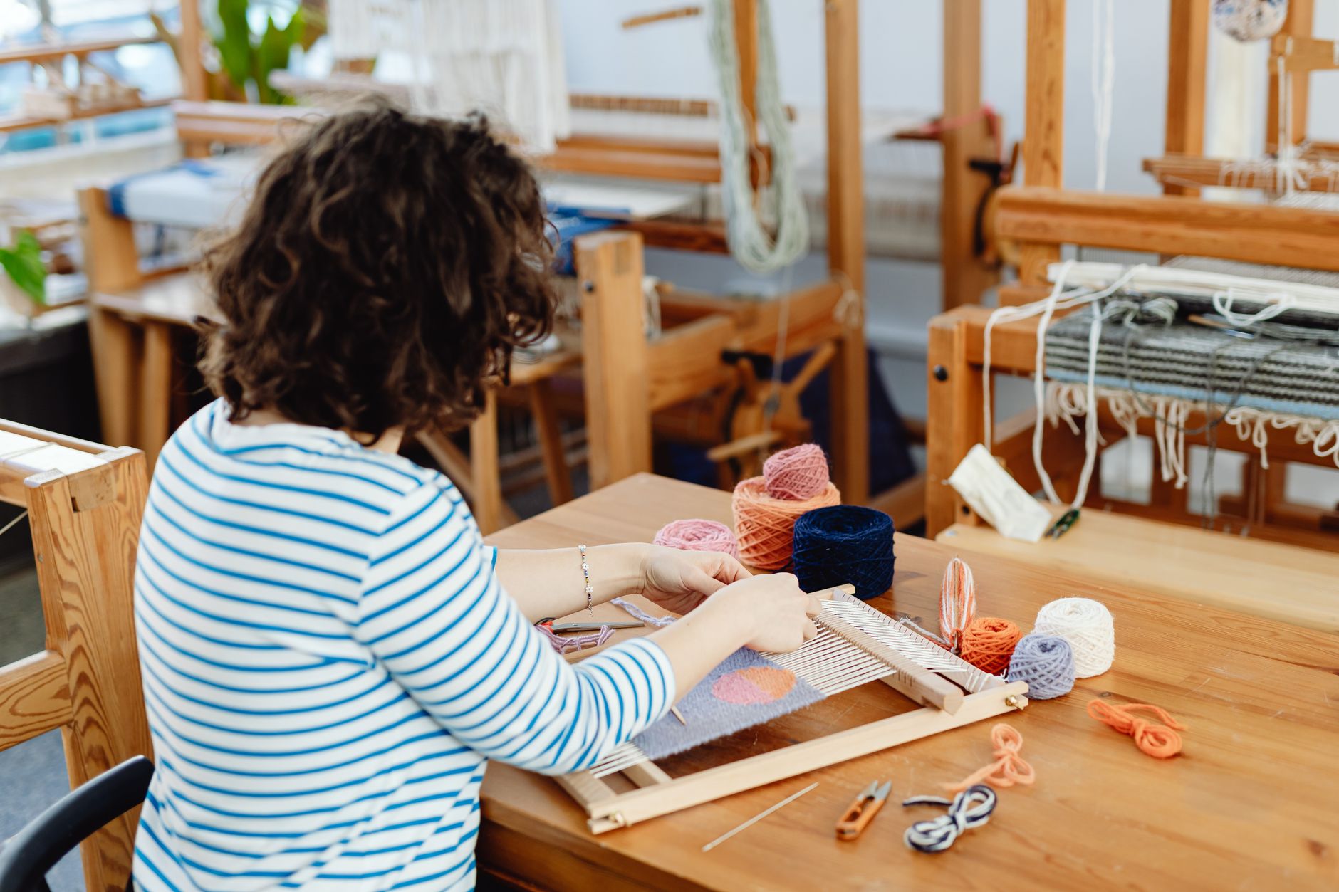 A woman focused on weaving with colorful yarn on a loom in a cozy indoor craft workshop.