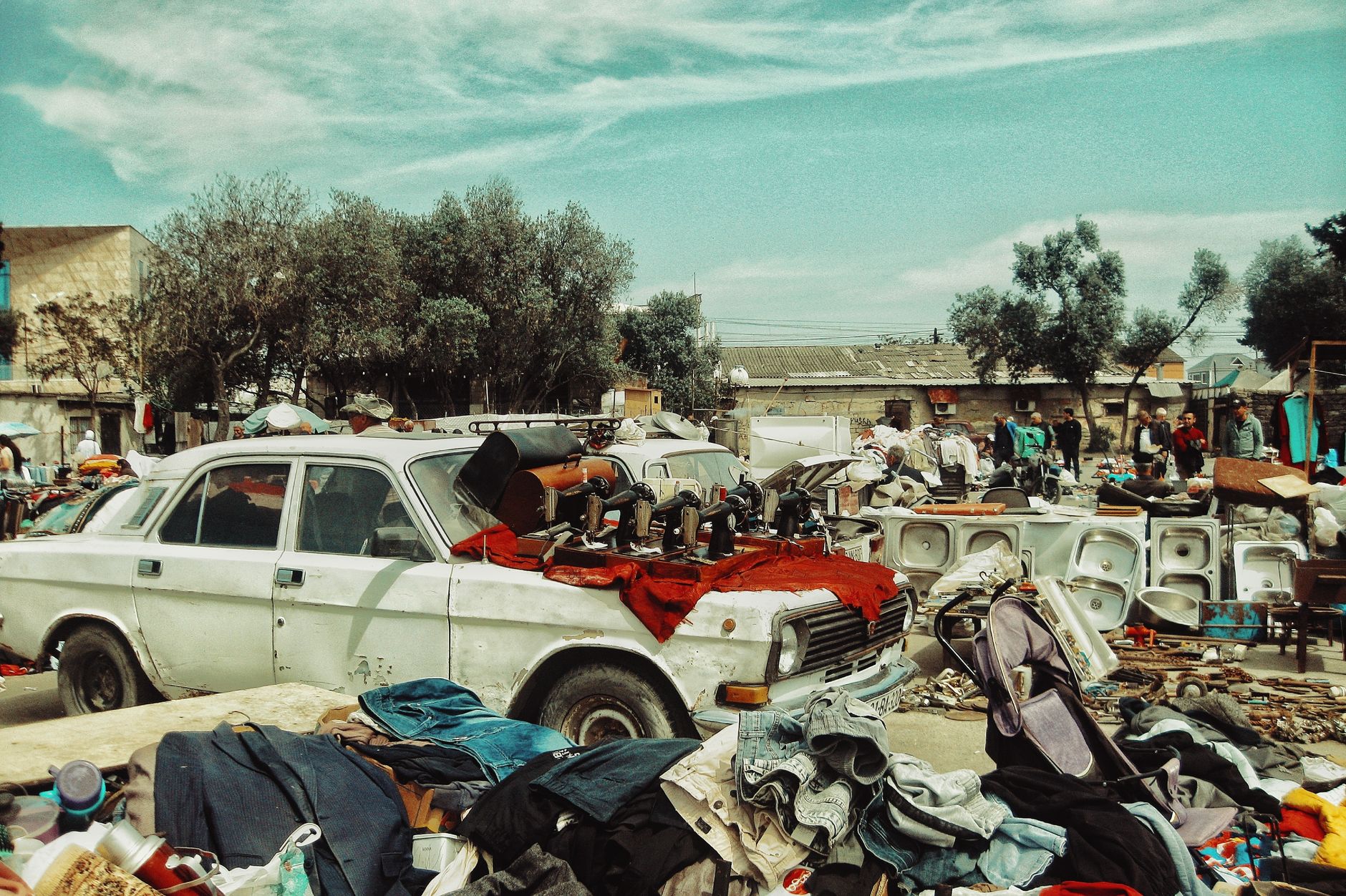 An outdoor flea market scene with a vintage car, sewing machines, clothes, and various items.