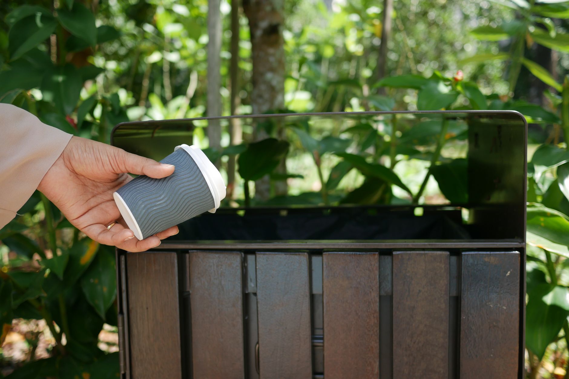A person throws a biodegradable coffee cup into a recycling bin amidst greenery.