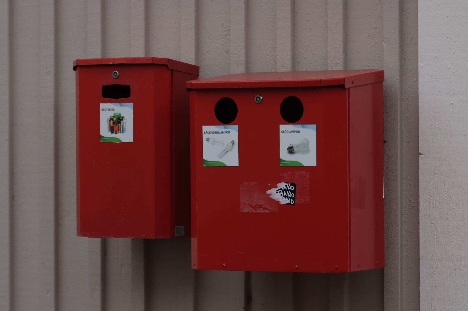 Two red recycling bins for batteries and light bulbs mounted on urban wall.