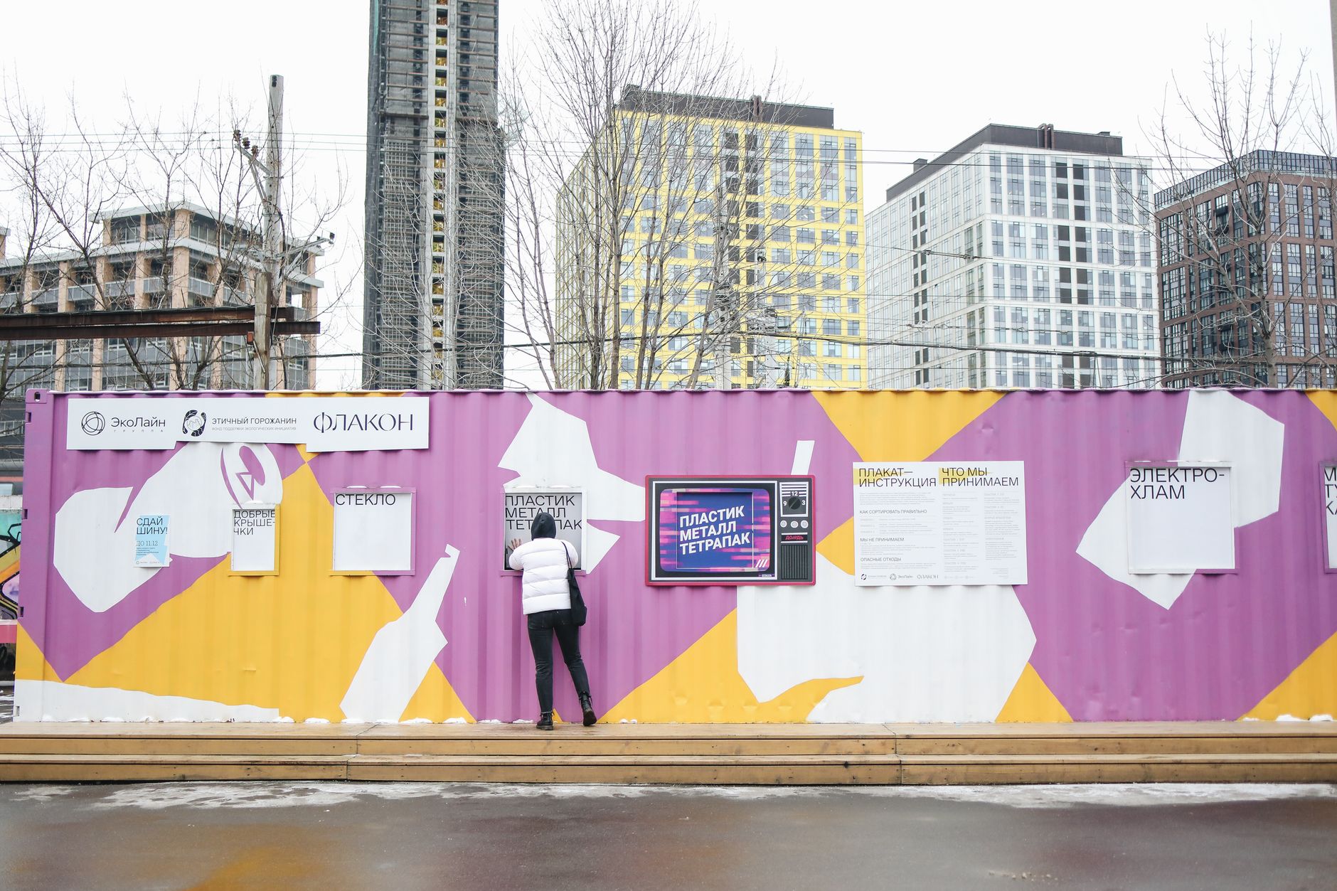 A person depositing waste at a vibrant urban recycling station with colorful containers and city buildings in background.