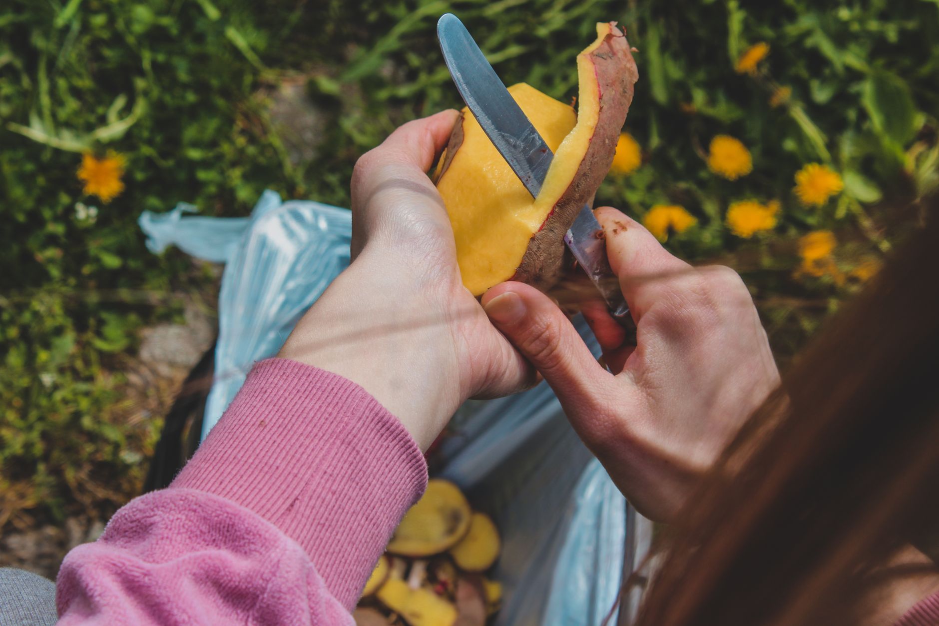 Hands peeling potatoes with a knife outdoors, showcasing food preparation and fresh produce.