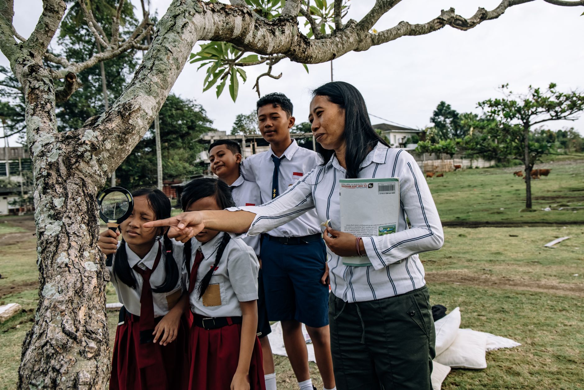 Students in school uniforms explore nature with their teacher using a magnifying glass.
