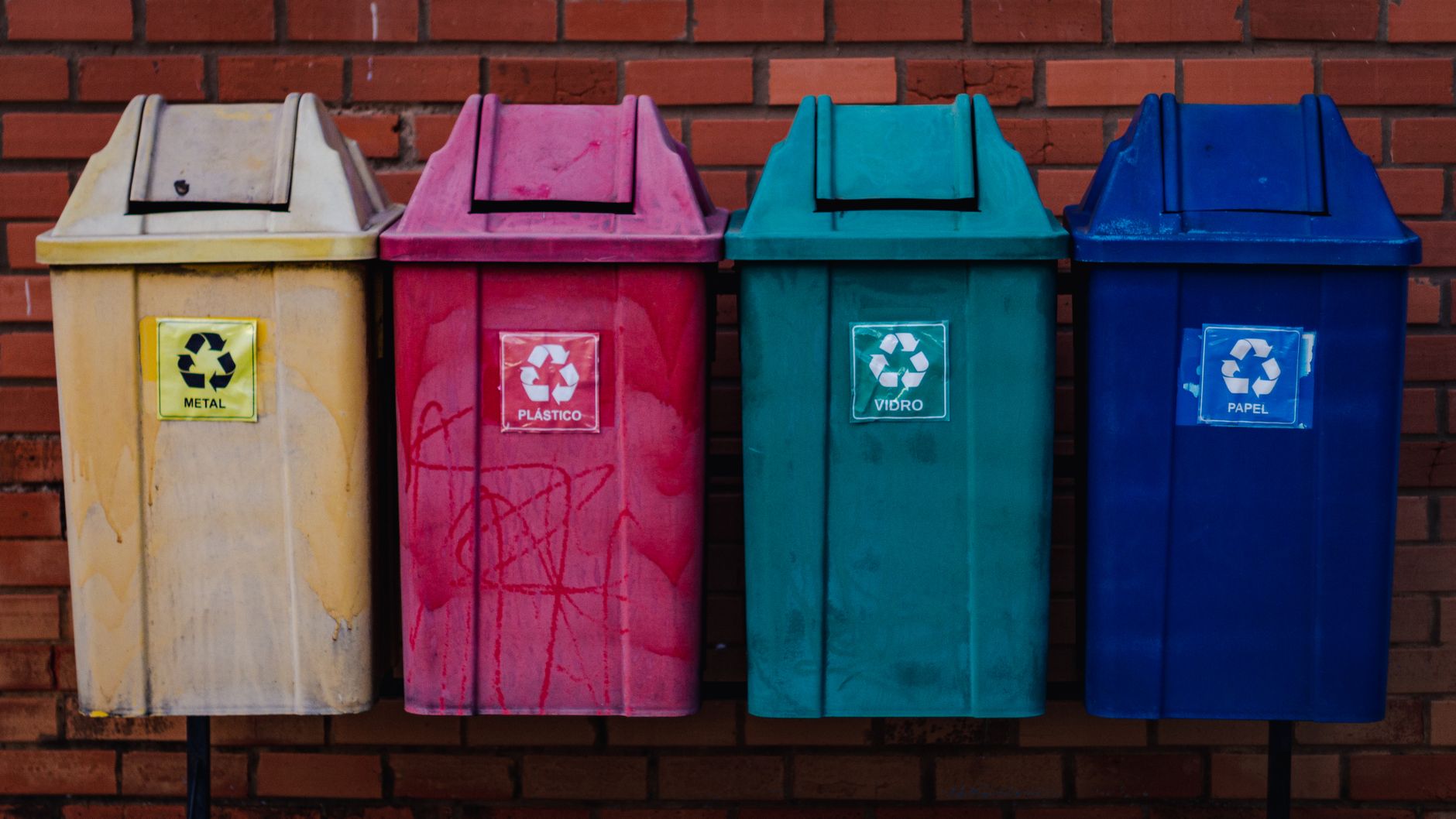 Four colorful recycling bins for metal, plastic, glass, and paper against a brick wall in Brazil.