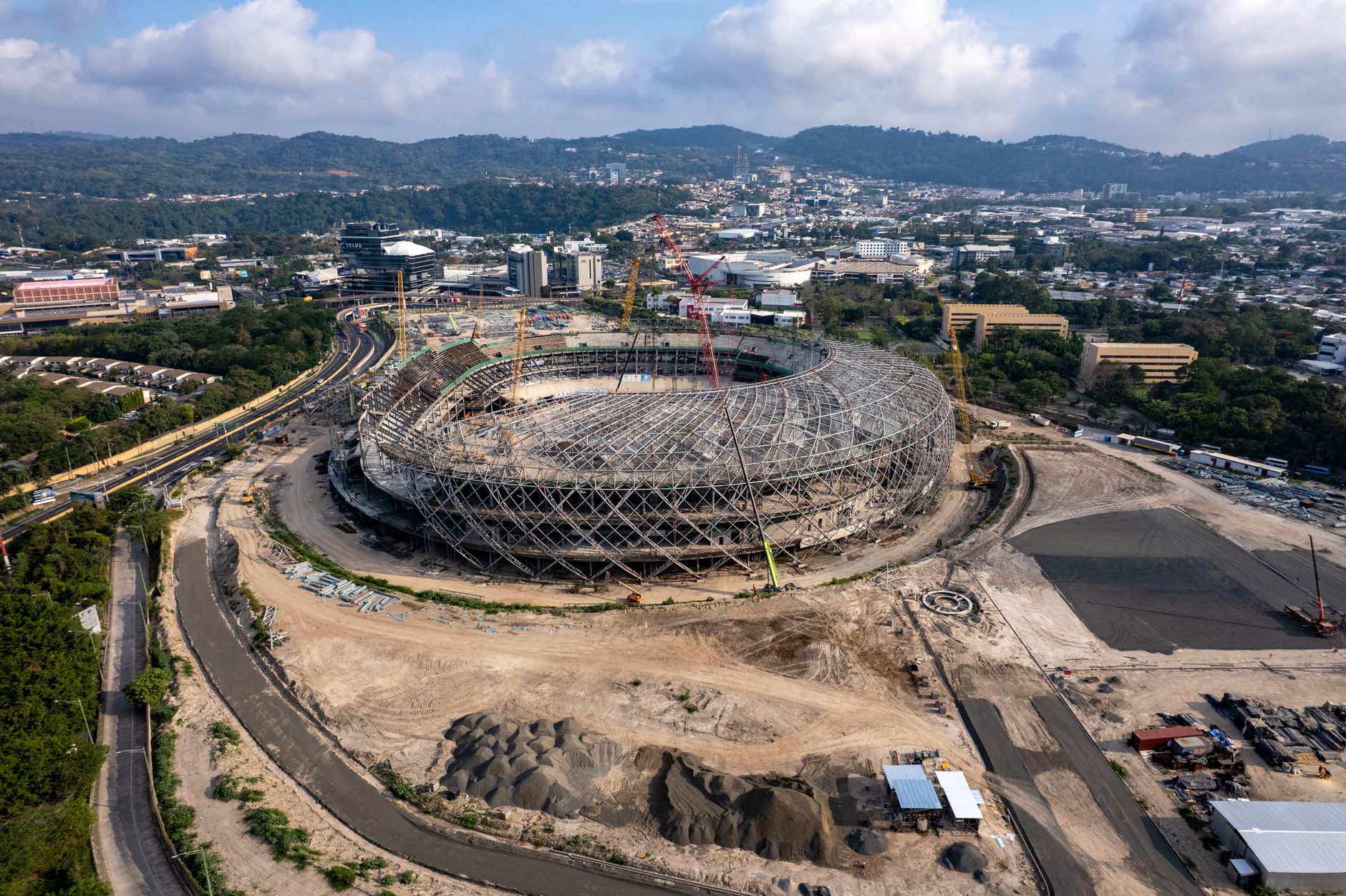 Aerial view of a large stadium under construction surrounded by urban areas and hills.