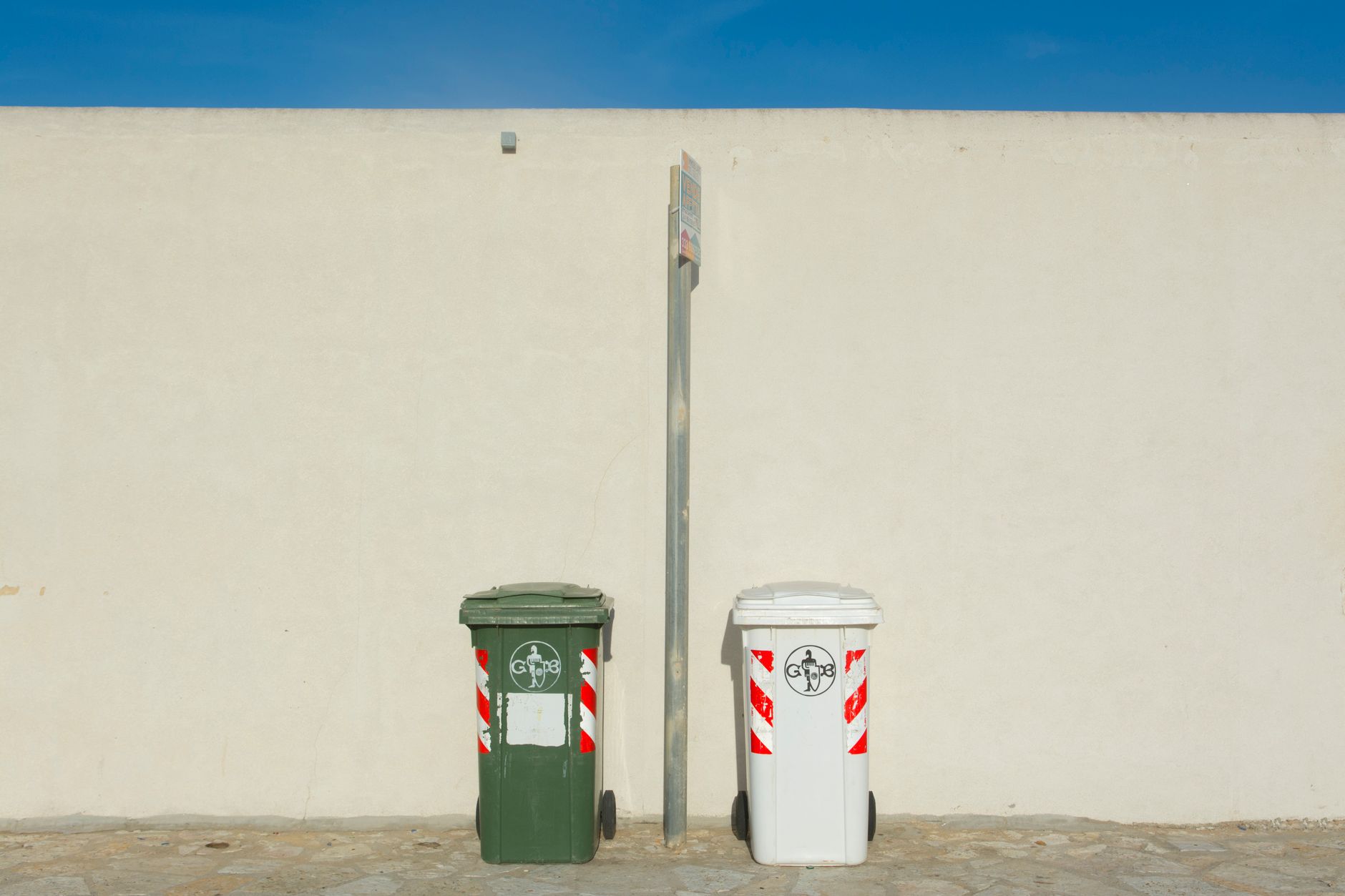 Two recycling bins next to a white wall under a clear blue sky, capturing urban simplicity.