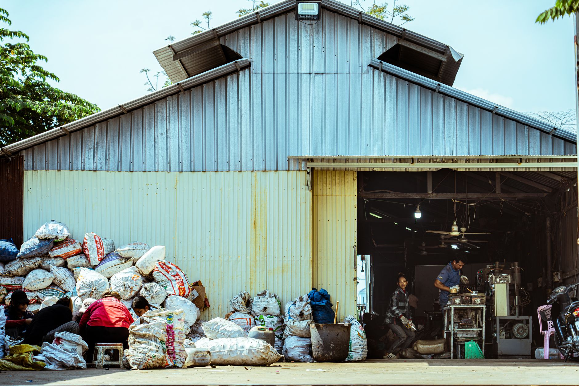 Industrial workers sorting recyclable materials outside a metal warehouse. Daytime scene showing recycling process.